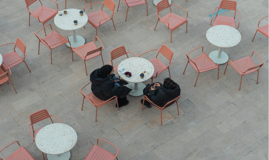 Aufnahme von oben auf die Terrasse eines Cafés mit vielen rosa Metallstühlen und weiß gesprenkelten Tischen, an einem Tisch sitzen zwei Personen in schwarzen Jacken.
